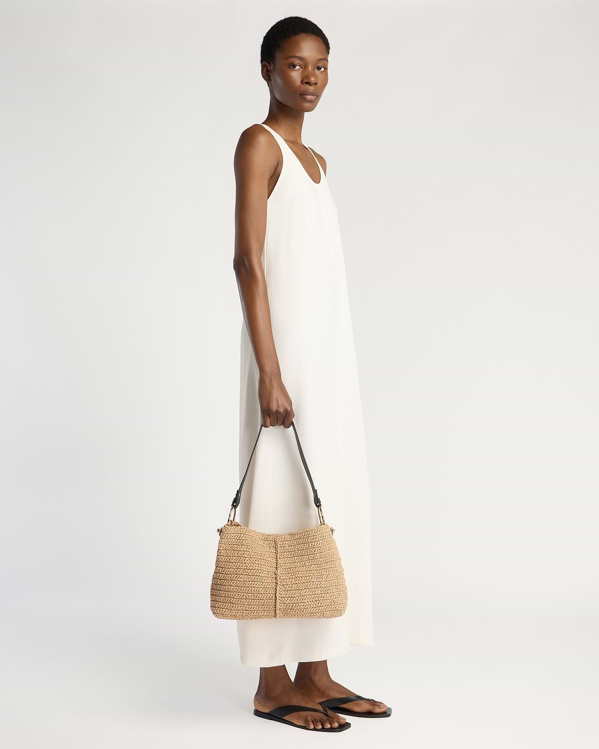 Model in white sleeveless dress holding raffia hobo bag with black strap