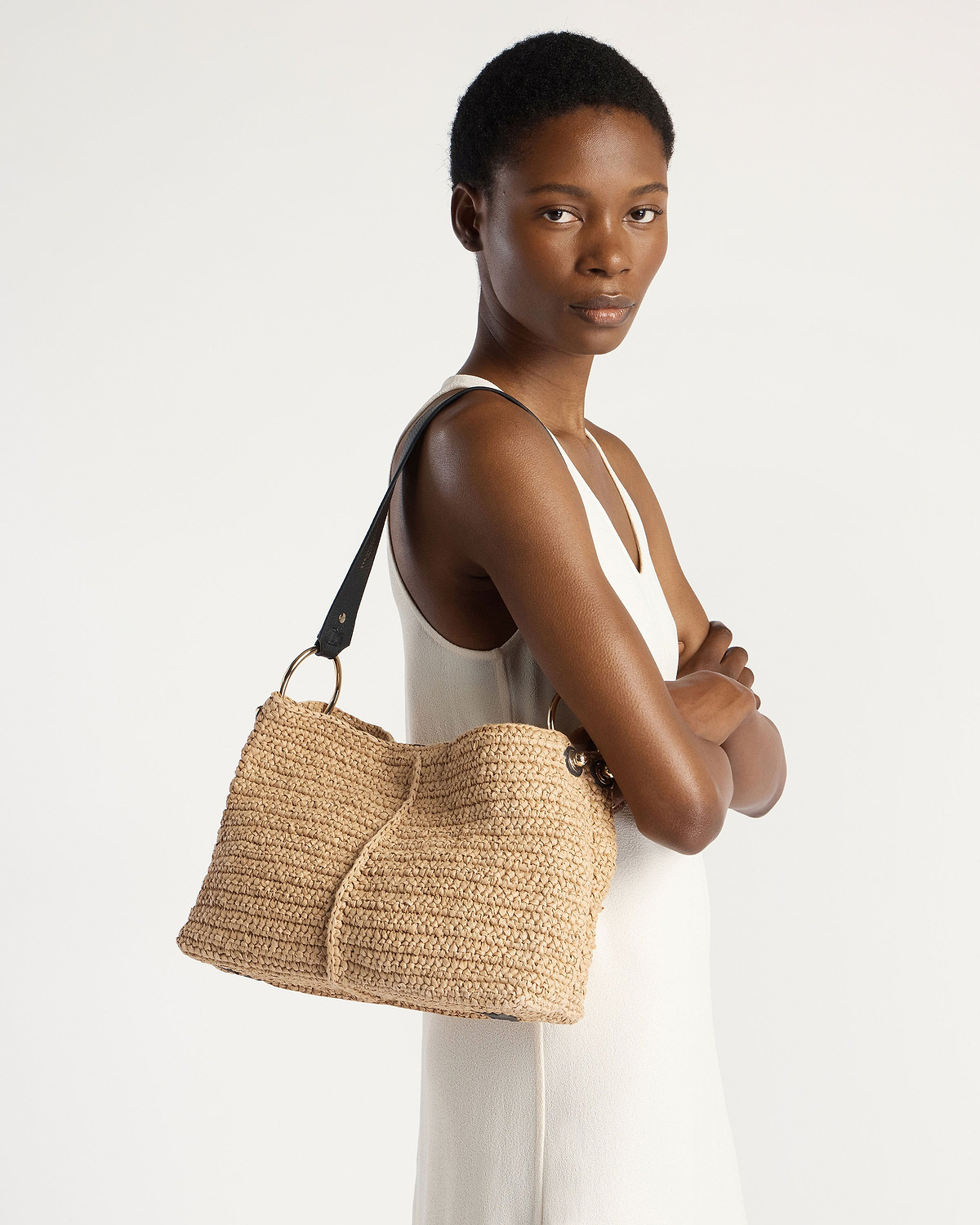 Model wearing white sleeveless dress carrying raffia hobo bag with black strap.