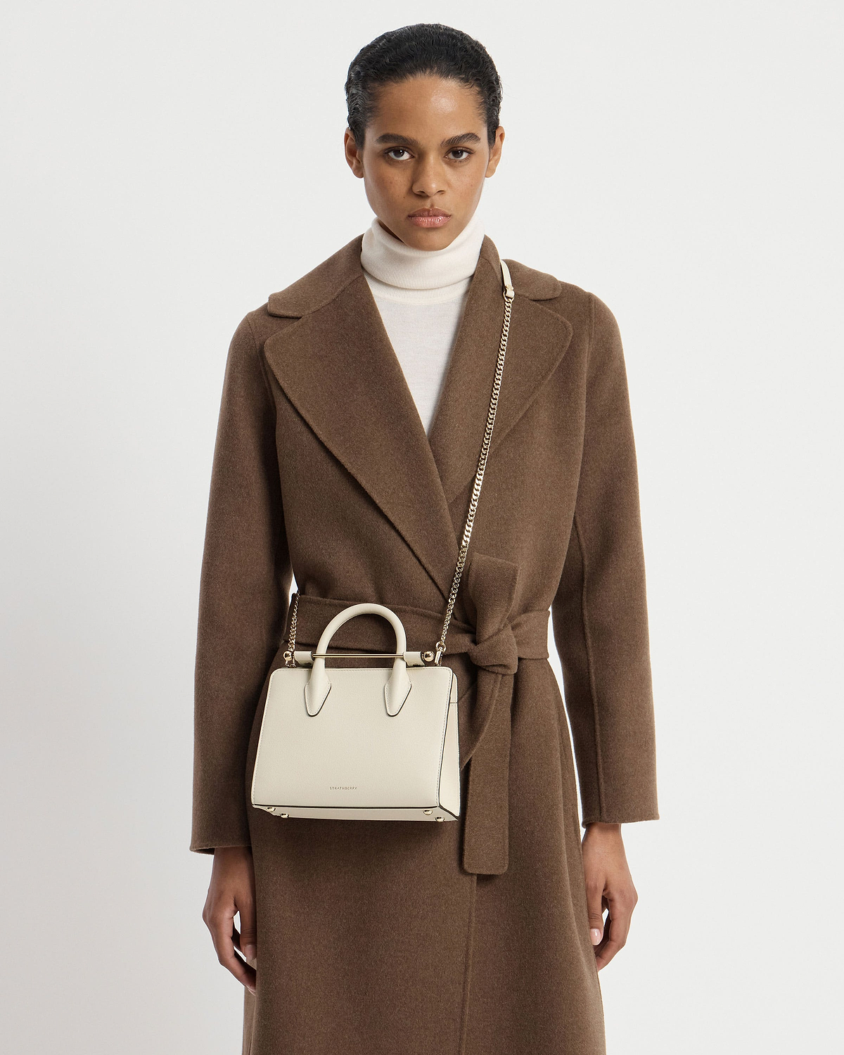 Model wearing brown coat with vanilla Strathberry mini tote bag on shoulder.
