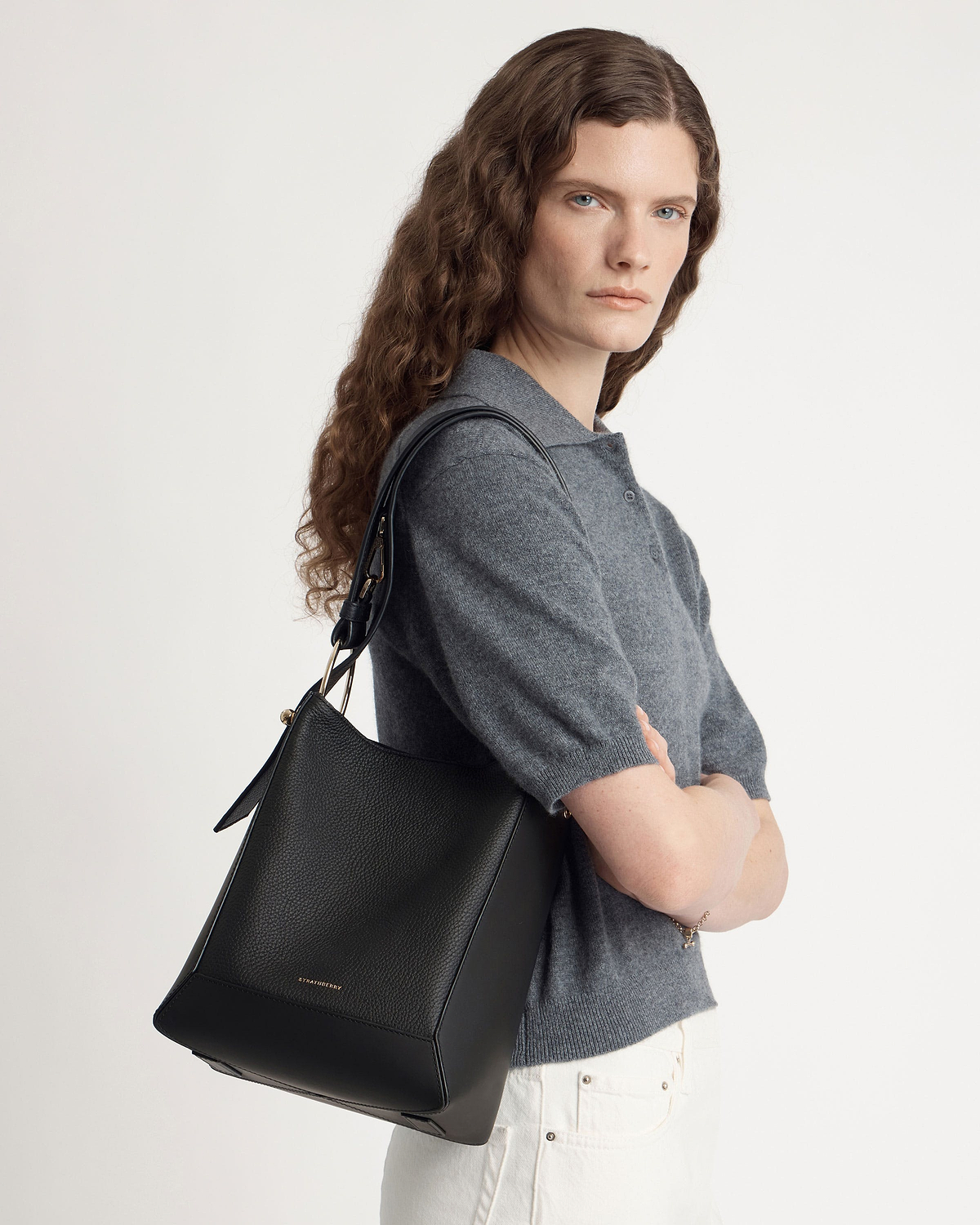 Woman wearing grey top carrying black Strathberry leather bucket bag on shoulder.