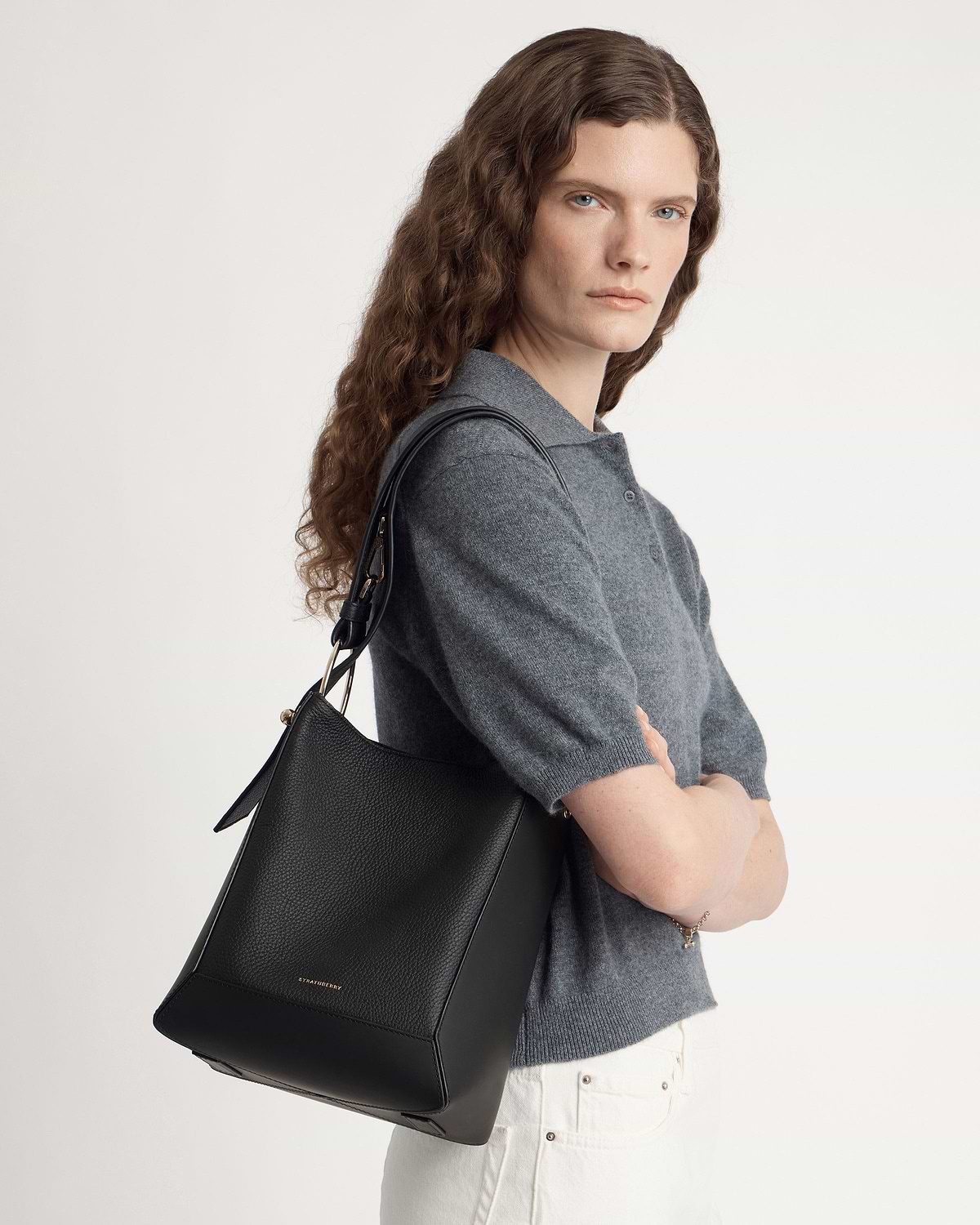 Woman wearing grey top carrying black Strathberry leather bucket bag on shoulder.