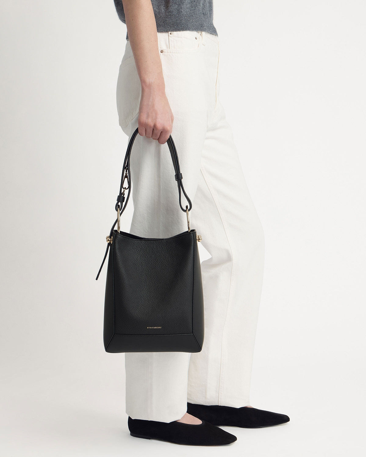 Model holding black Strathberry bucket bag with gold hardware and white trousers.