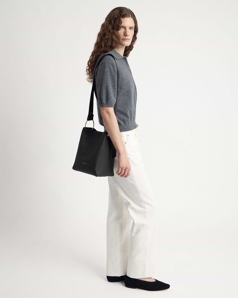 Model wearing grey short-sleeve polo and white jeans carrying black bucket bag over shoulder