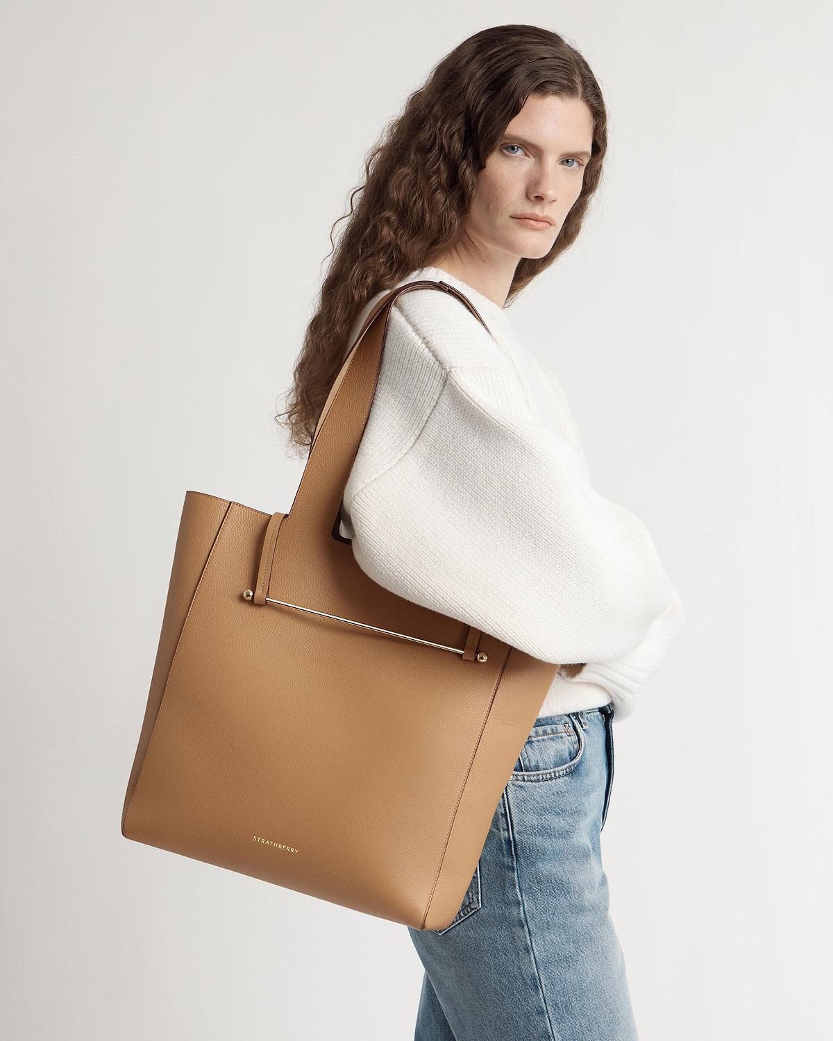 Woman in white jumper and jeans carrying Strathberry caramel tote on shoulder.