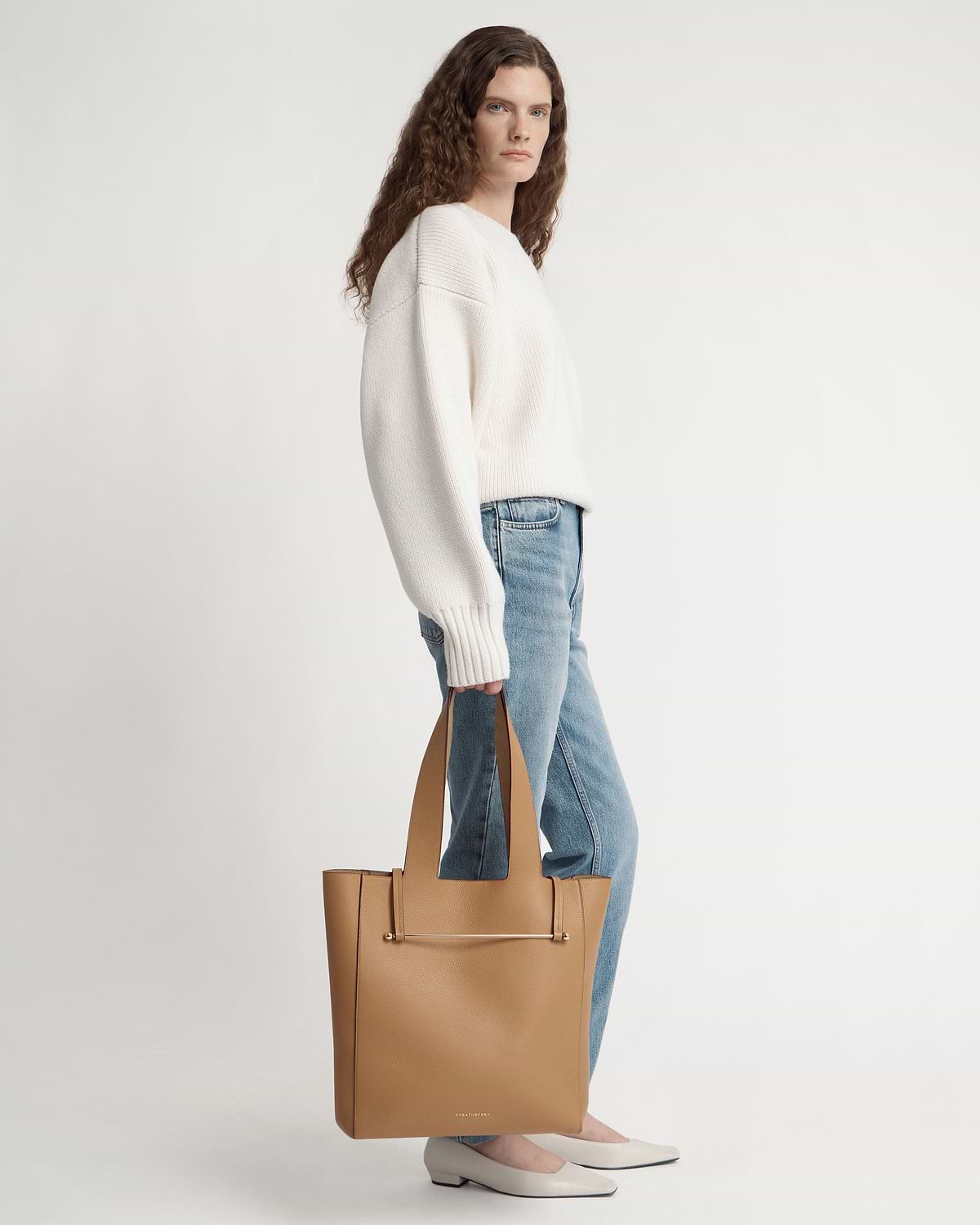 Woman standing in white jumper and jeans holding a caramel tote bag