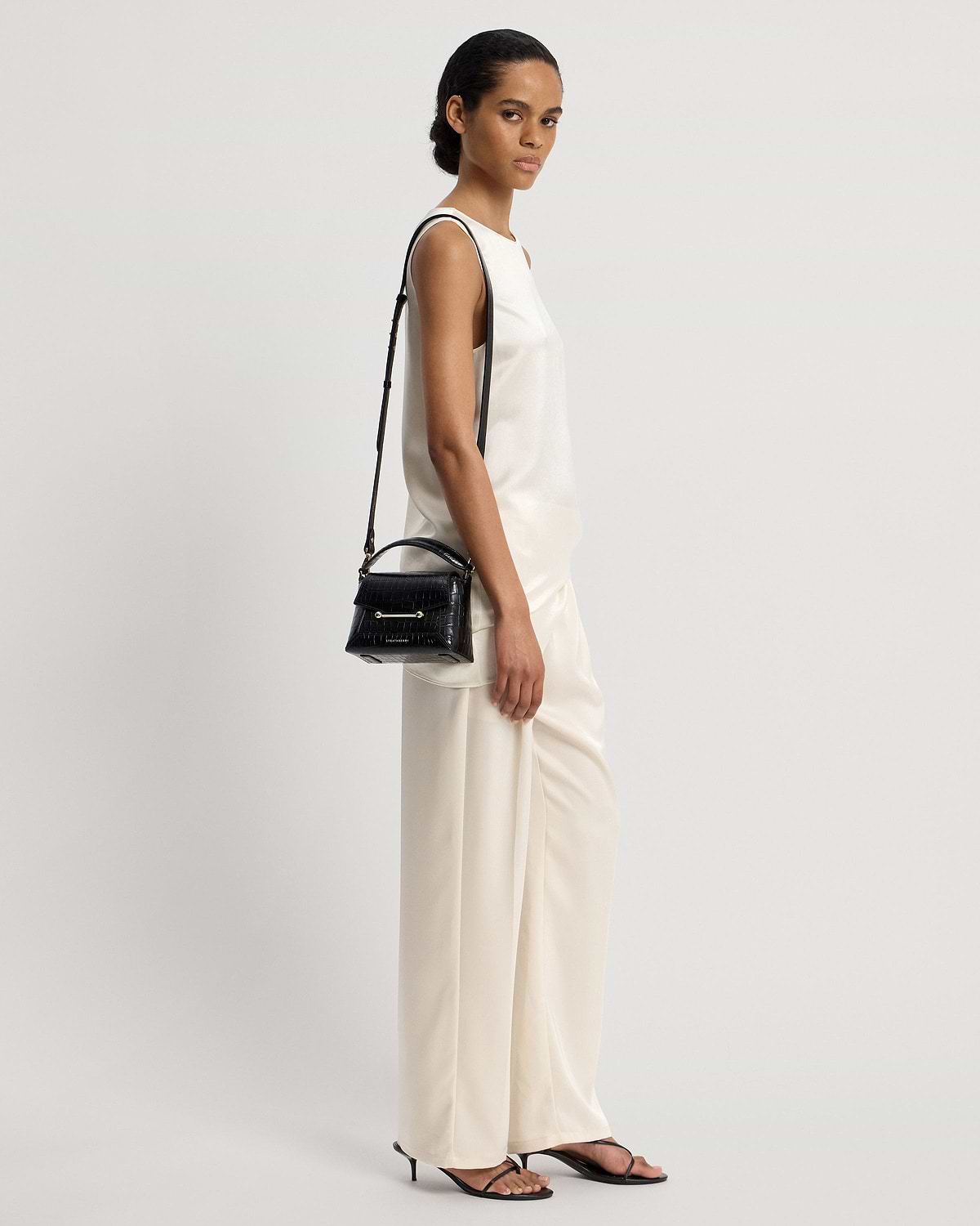 Model wearing cream outfit carrying small black croc-embossed top-handle bag with shoulder strap.
