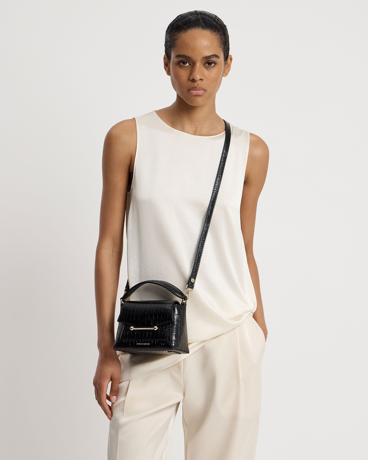 Model wearing cream top and small black croc top-handle crossbody bag with Strathberry logo