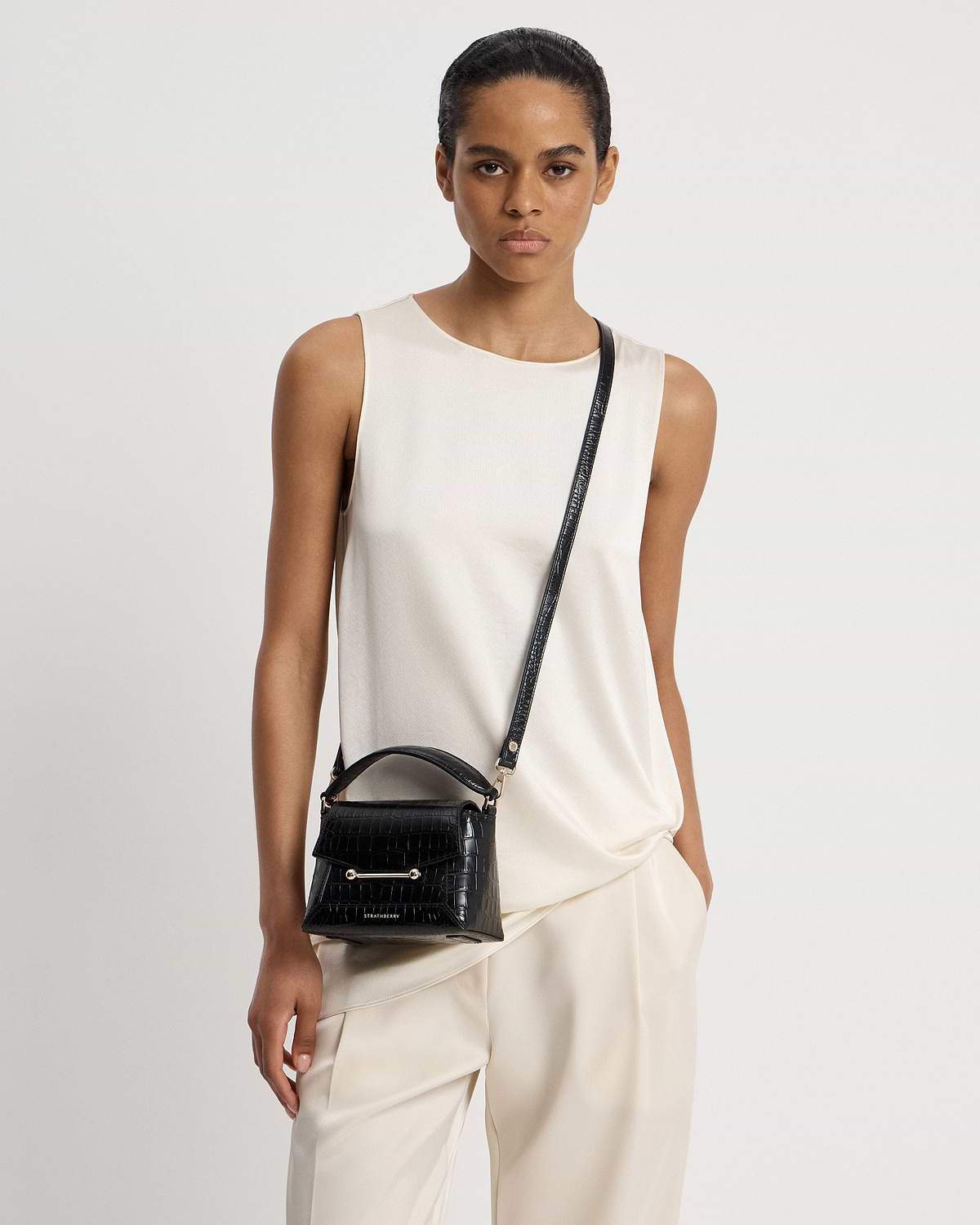 Model wearing cream top and small black croc top-handle crossbody bag with Strathberry logo