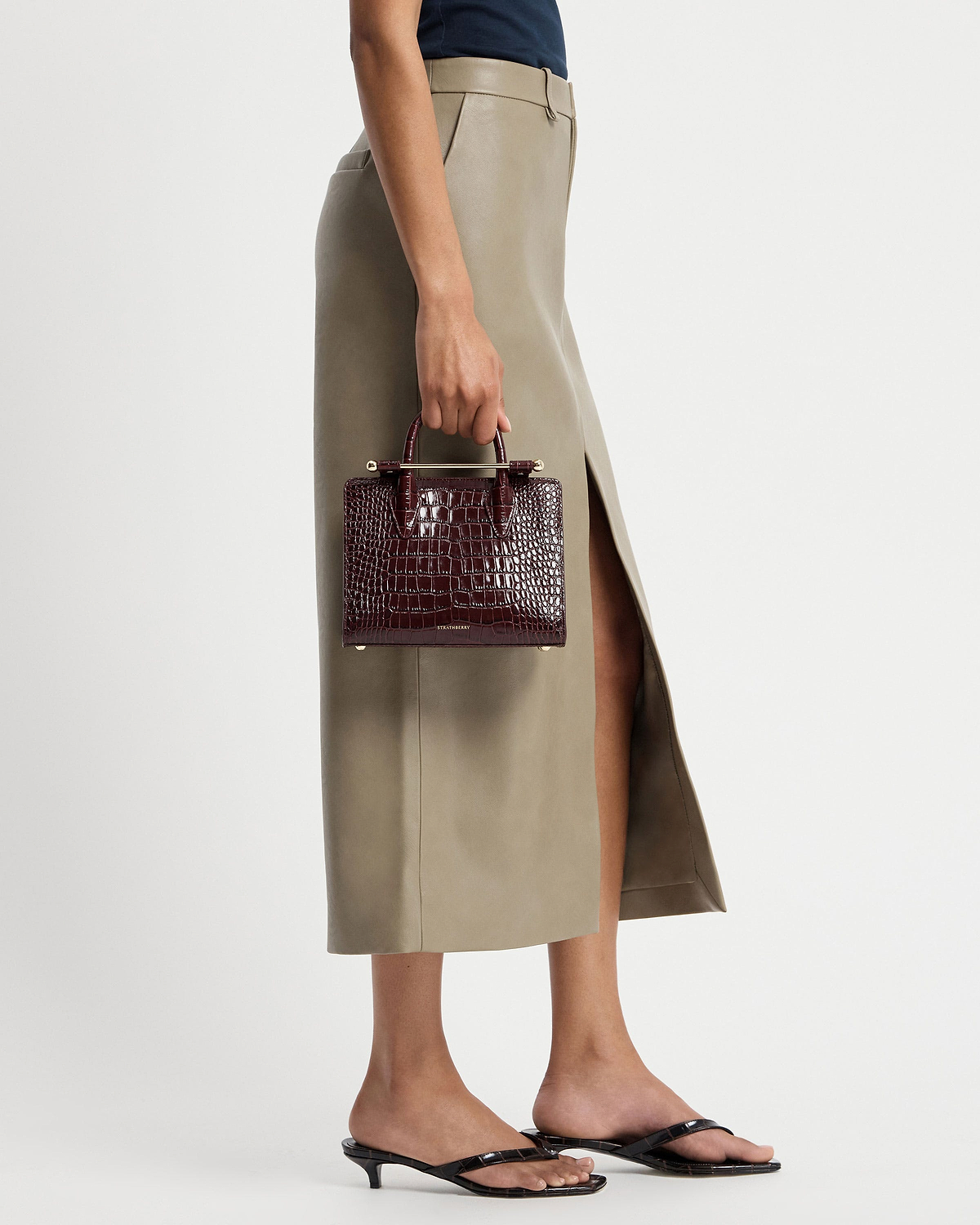 Model's hand holding burgundy croc mini tote by Strathberry beside skirt.
