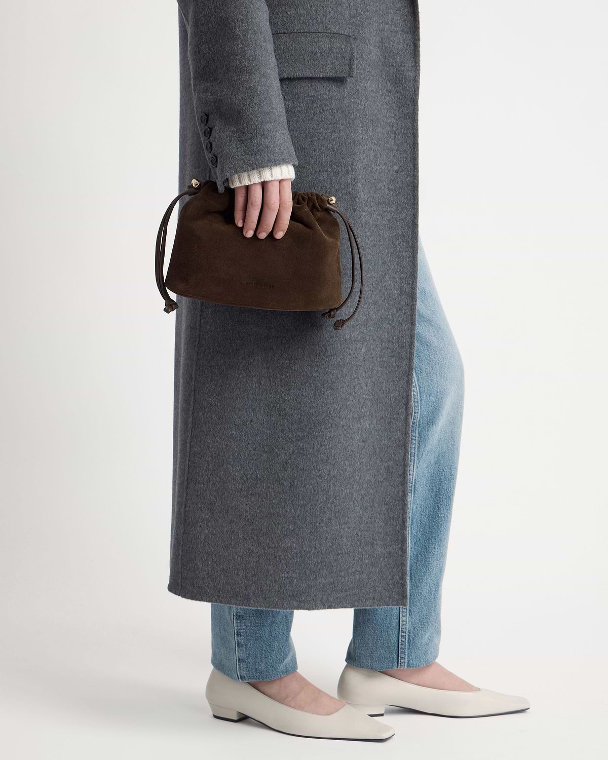 Model holding chocolate suede drawstring bag with grey coat, jeans, cream flats.