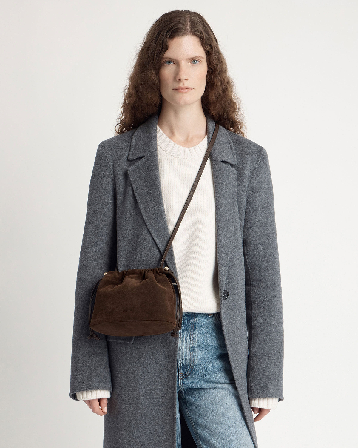 Model wearing a grey coat and jeans with a chocolate suede crossbody drawstring bag.