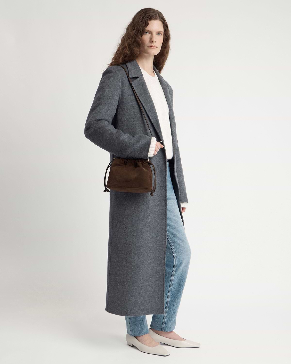 Model wearing long grey coat and jeans carrying chocolate suede drawstring bag.