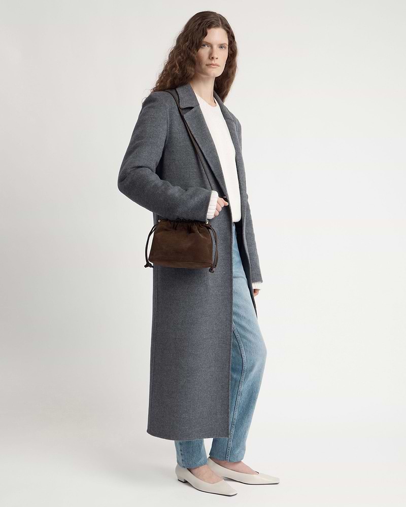 Model wearing long grey coat and jeans carrying chocolate suede drawstring bag.