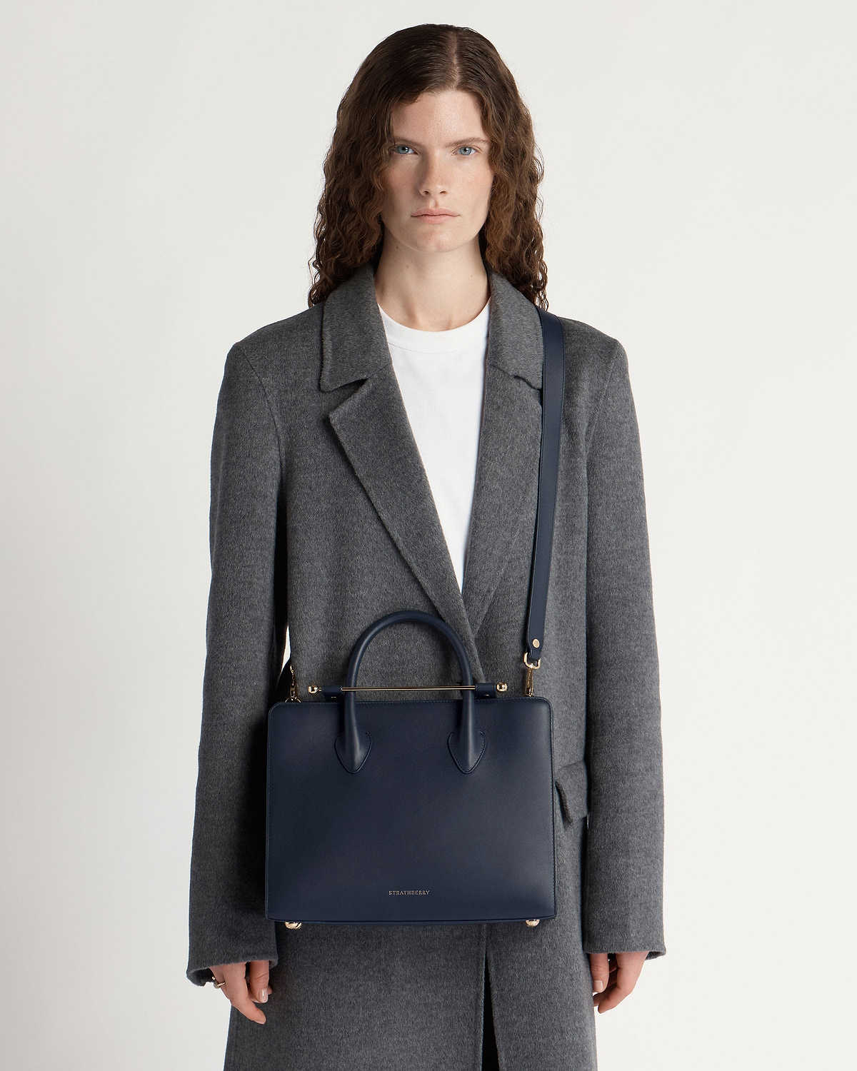 Model in grey coat carrying marine blue Strathberry midi tote with shoulder strap.