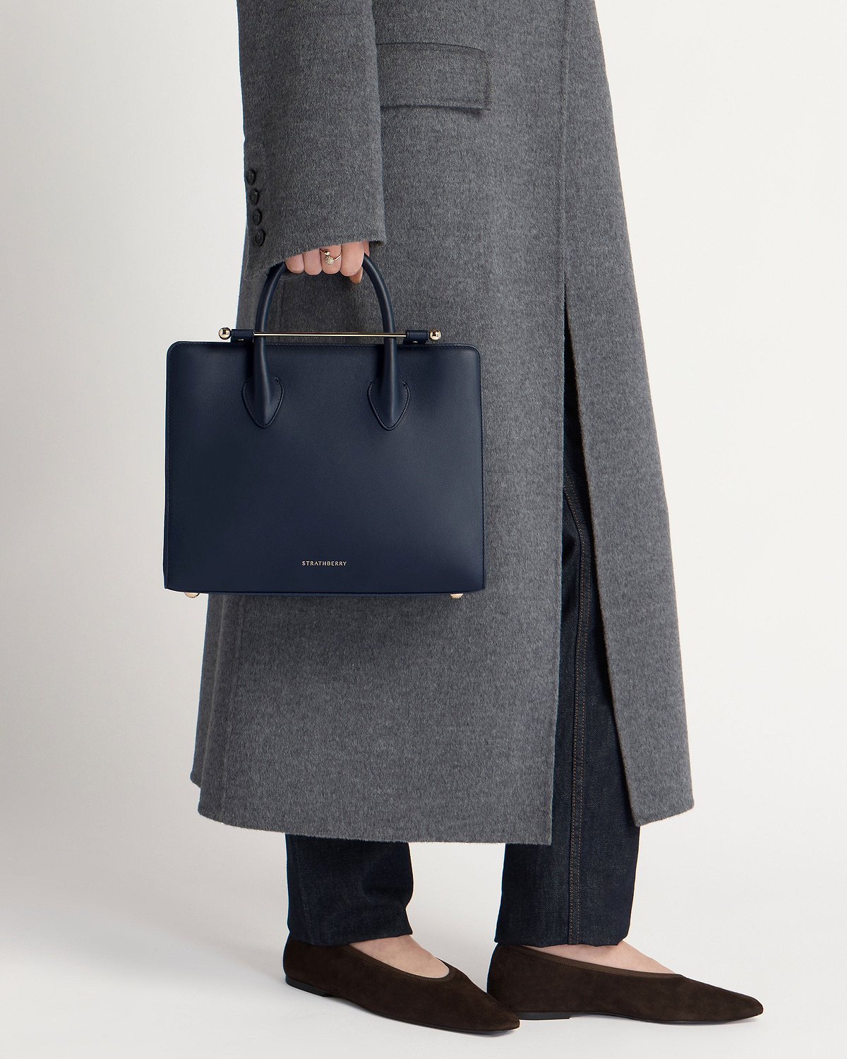 A hand holds Strathberry marine blue midi tote against grey coat and jeans.