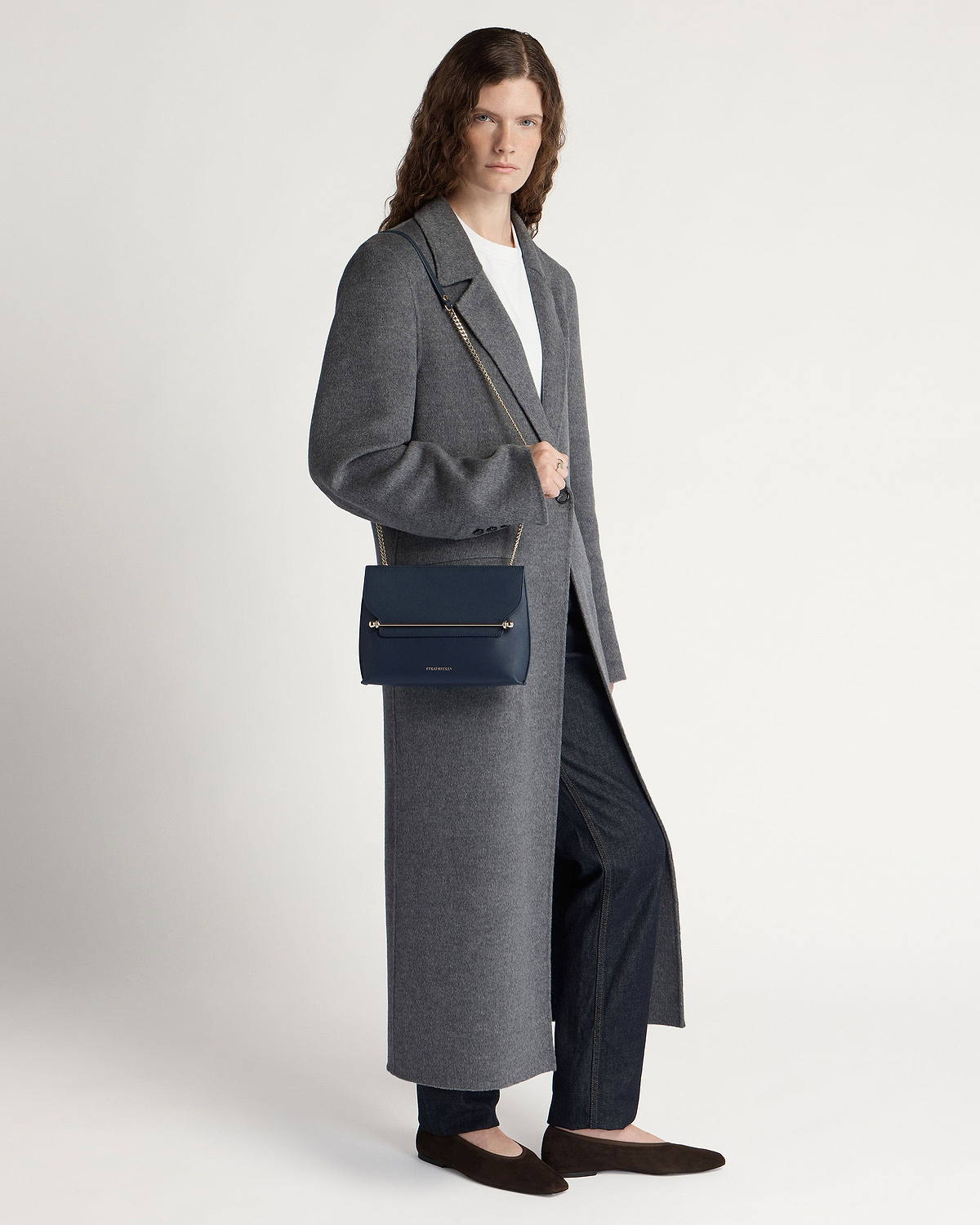 Model wearing long grey coat carrying marine blue shoulder bag with gold bar detail.