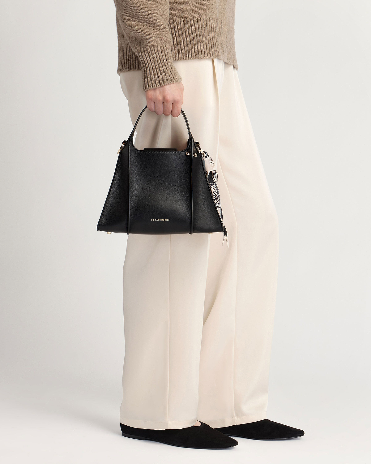 Model holding black Strathberry Arkshell handbag with patterned scarf, cream trousers.