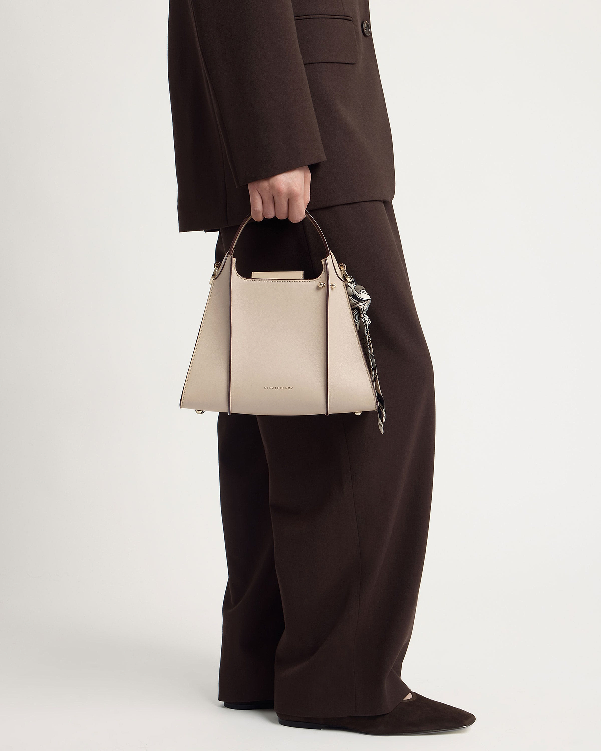 Model holding Strathberry Arkshell oat handbag by its top handle next to brown suit trousers.