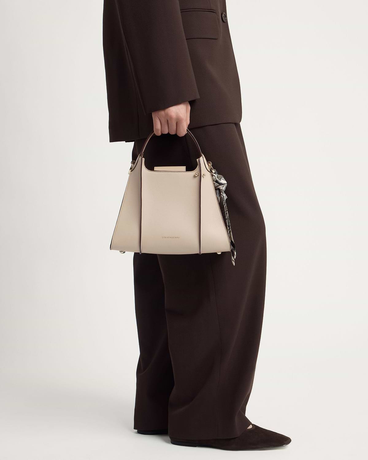 Model holding Strathberry Arkshell oat handbag by its top handle next to brown suit trousers.