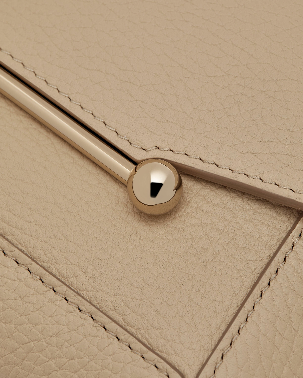 Close up of sand leather flap showing stitching, a gold bar and round stud.