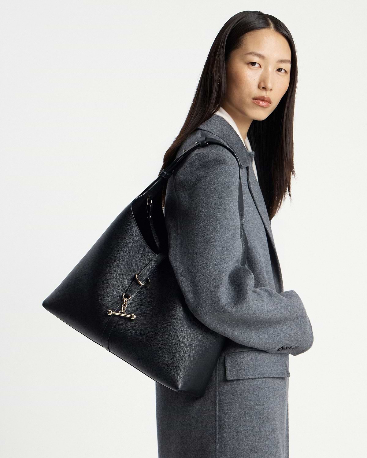 Model wearing a grey coat carrying a black hobo shoulder bag with gold hardware.