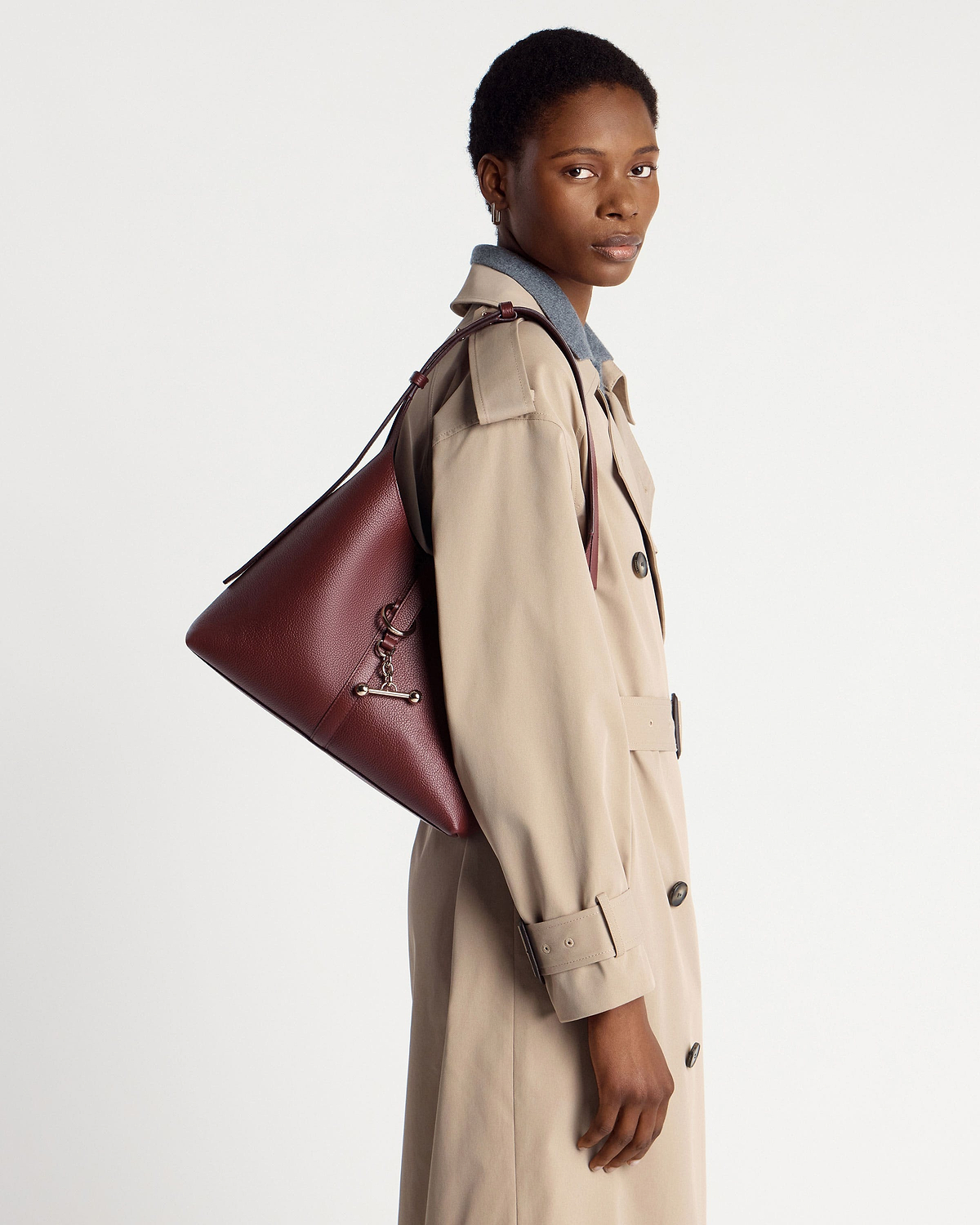 Model wearing beige trench coat carrying oxblood leather hobo shoulder bag.