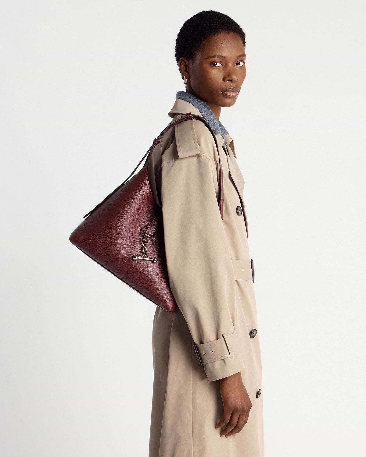 Model wearing beige trench coat carrying oxblood leather hobo shoulder bag.