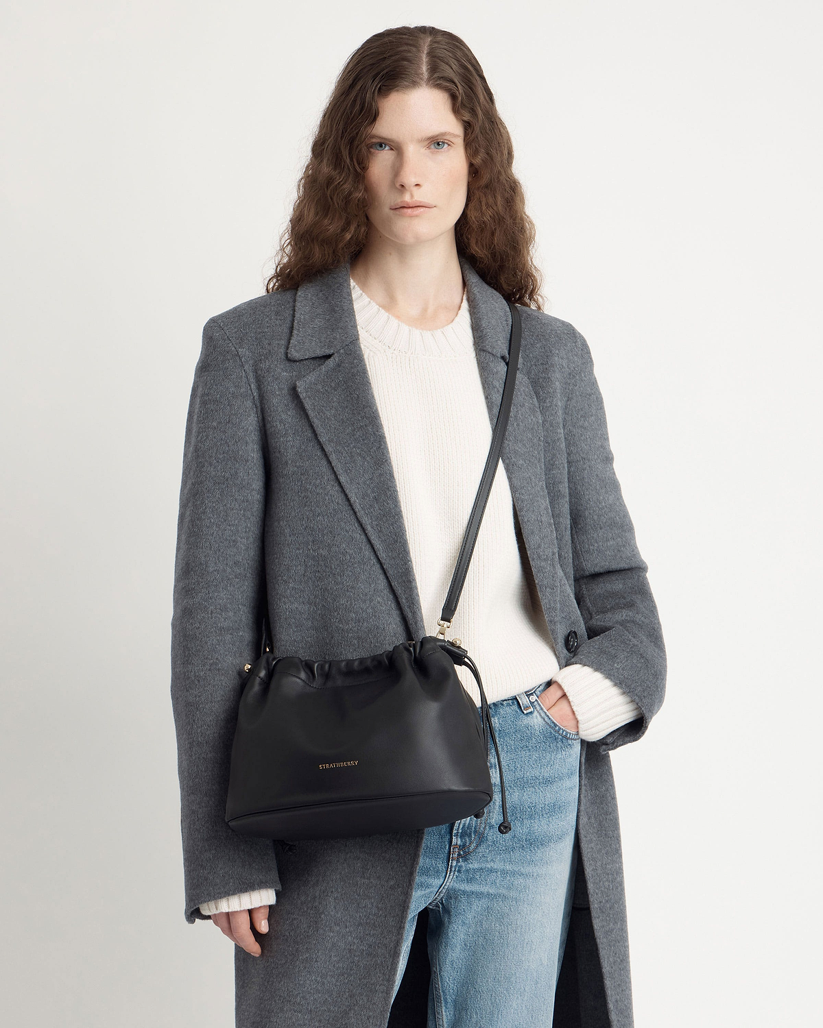 Model wearing grey coat and white sweater with black Strathberry crossbody drawstring bag.