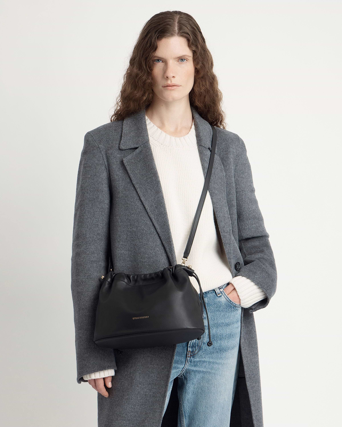 Model wearing grey coat and white sweater with black Strathberry crossbody drawstring bag.