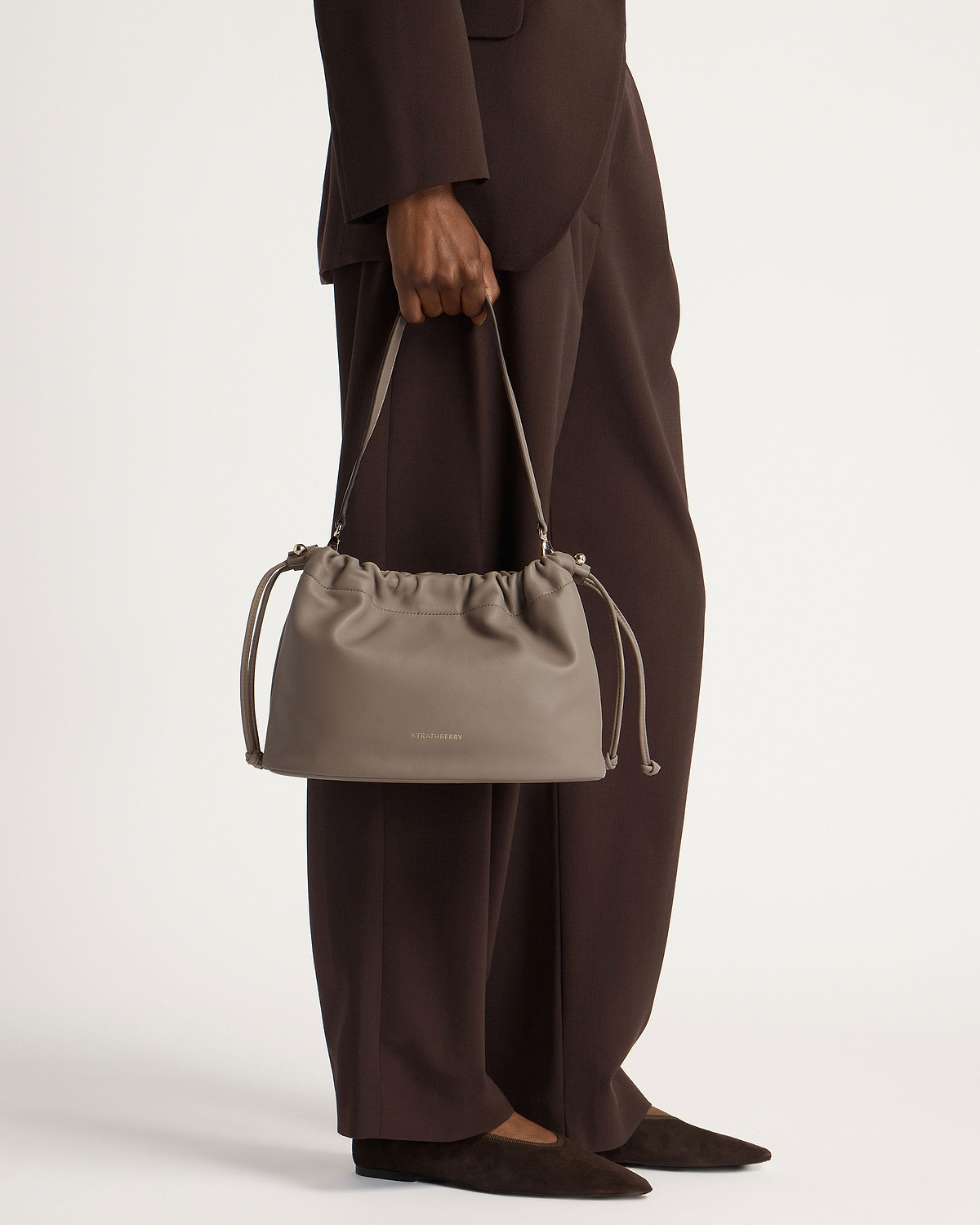 Model holding taupe Strathberry Charlotte Midi drawstring bag with shoulder handle