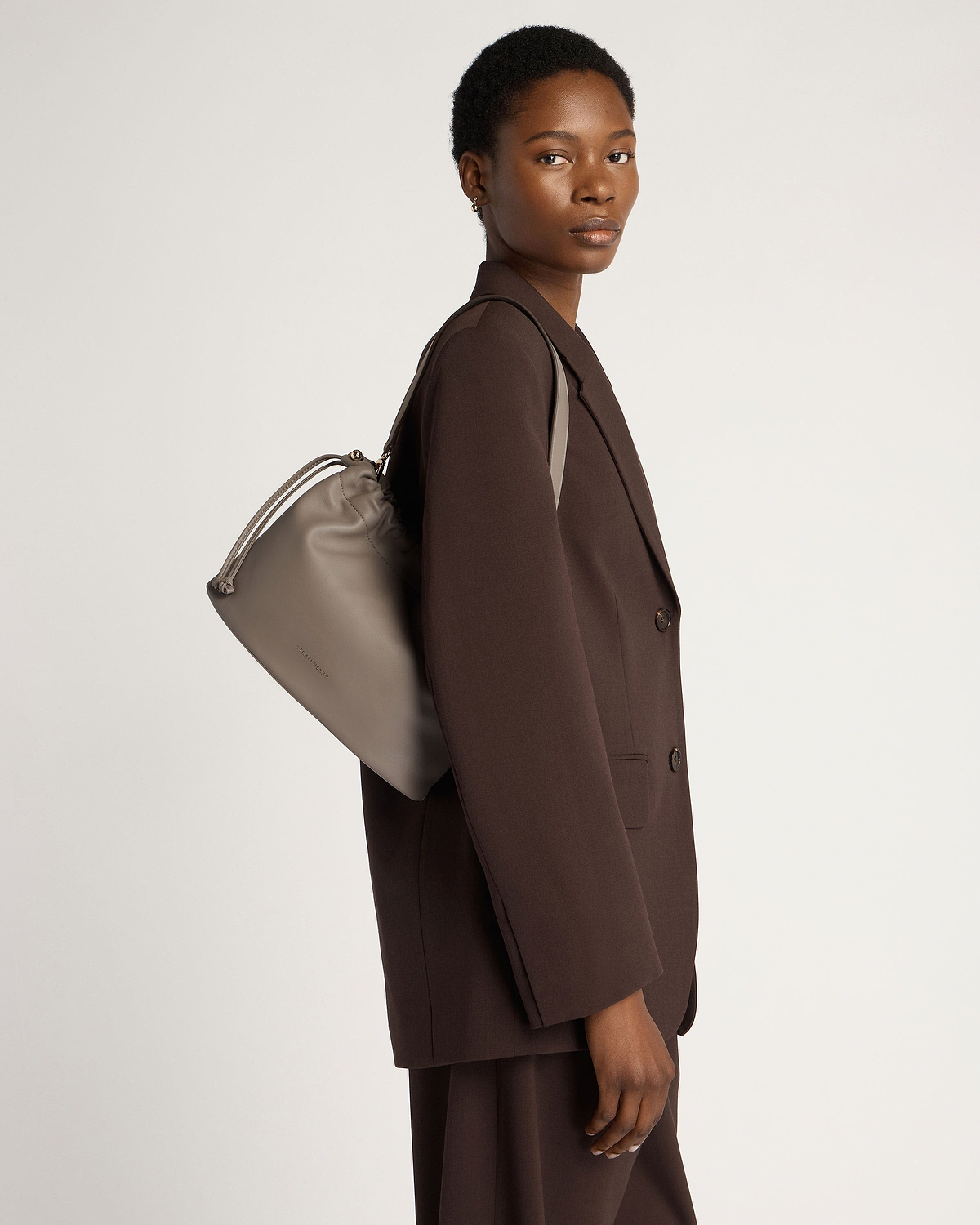 Model wearing a brown blazer carrying a taupe drawstring shoulder bag.