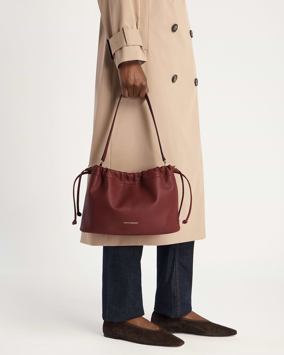 Model holds an oxblood Charlotte Midi drawstring bag with Strathberry logo.