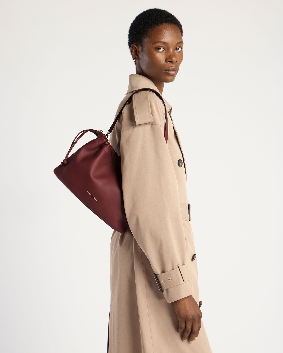 Model wearing a beige trench coat carrying oxblood Strathberry drawstring bag.