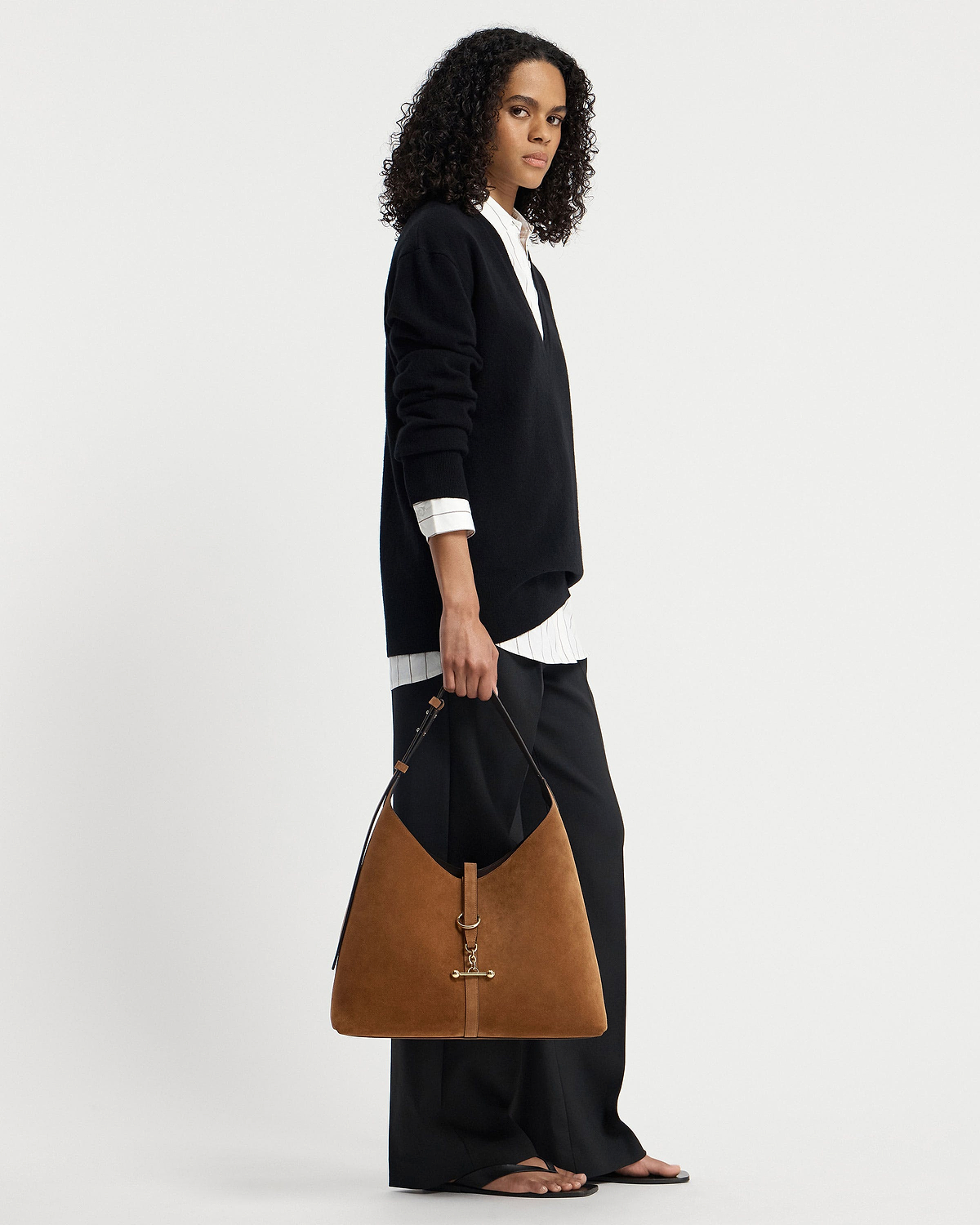 Model holding tan suede hobo bag with gold hardware, wearing black jumper.