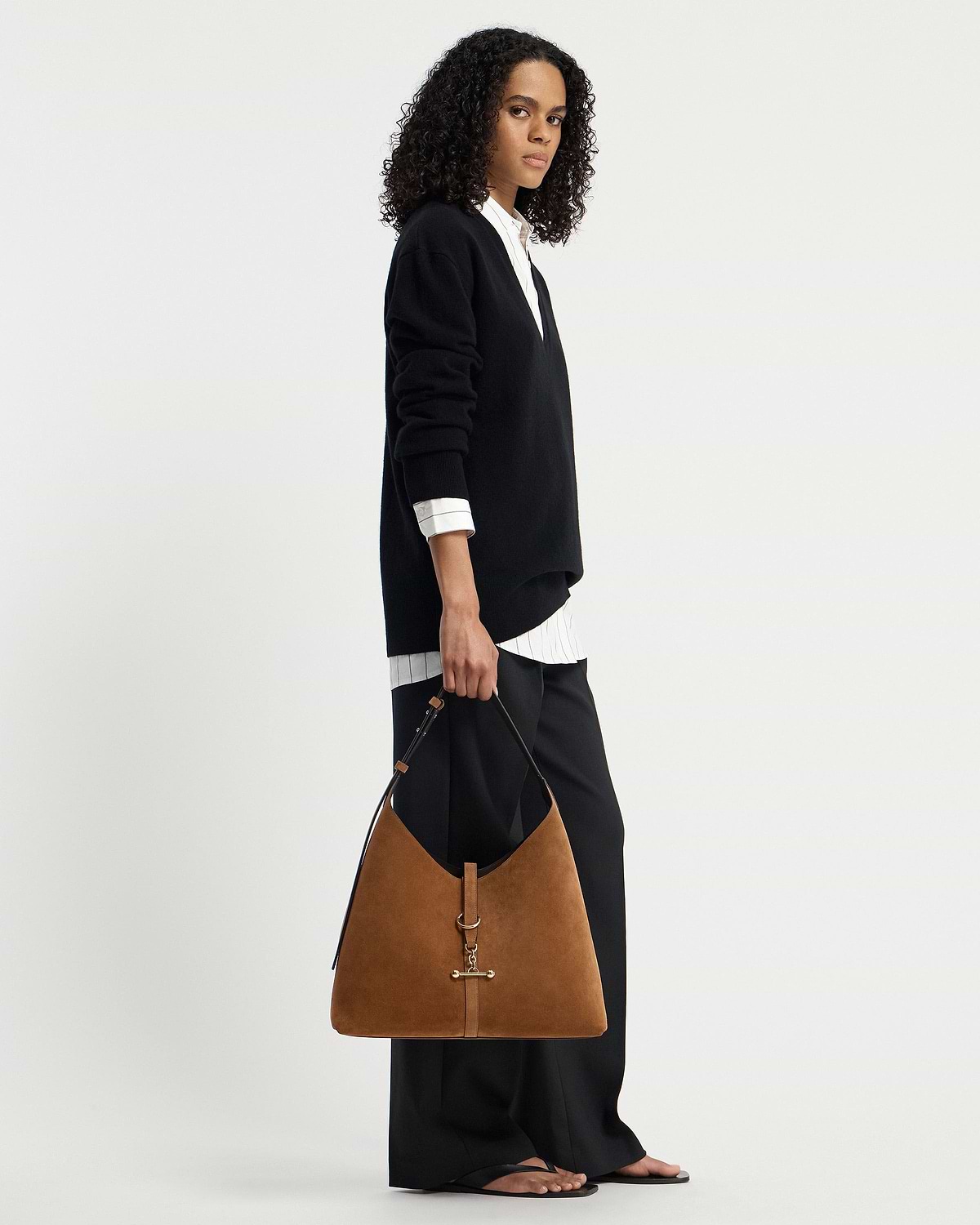 Model holding tan suede hobo bag with gold hardware, wearing black jumper.