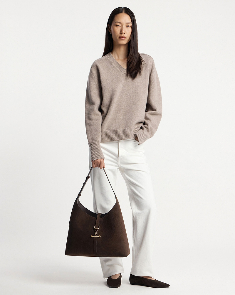 Model wearing beige V‑neck jumper and white trousers holding chocolate suede hobo bag.