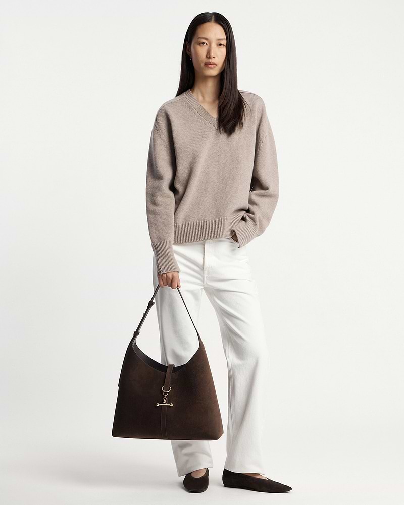 Model wearing beige V‑neck jumper and white trousers holding chocolate suede hobo bag.