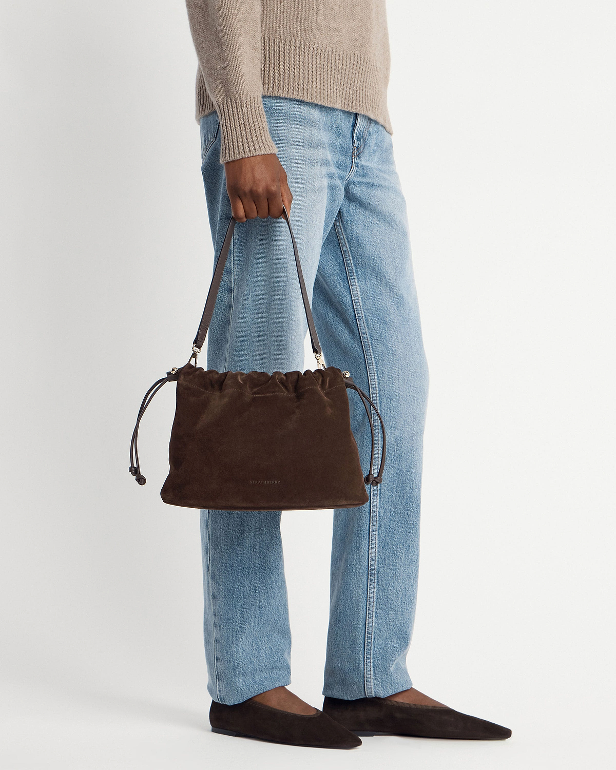 Chocolate suede Strathberry Charlotte midi drawstring bag held by model wearing jeans and flats.