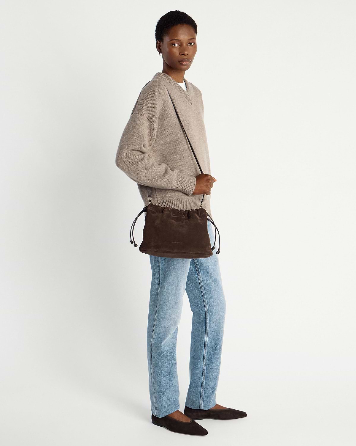 Model wearing chocolate suede drawstring bag crossbody, beige jumper and light jeans.