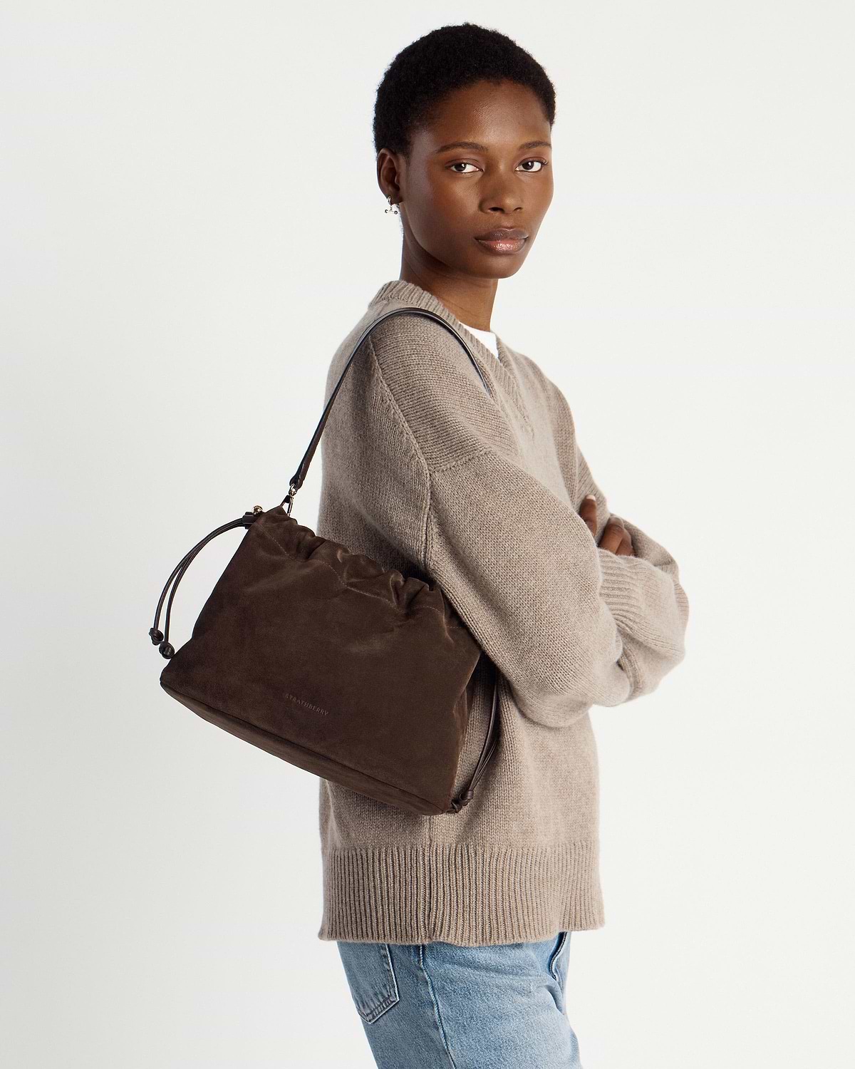 Model wearing a chocolate suede drawstring bag over shoulder, arms crossed.