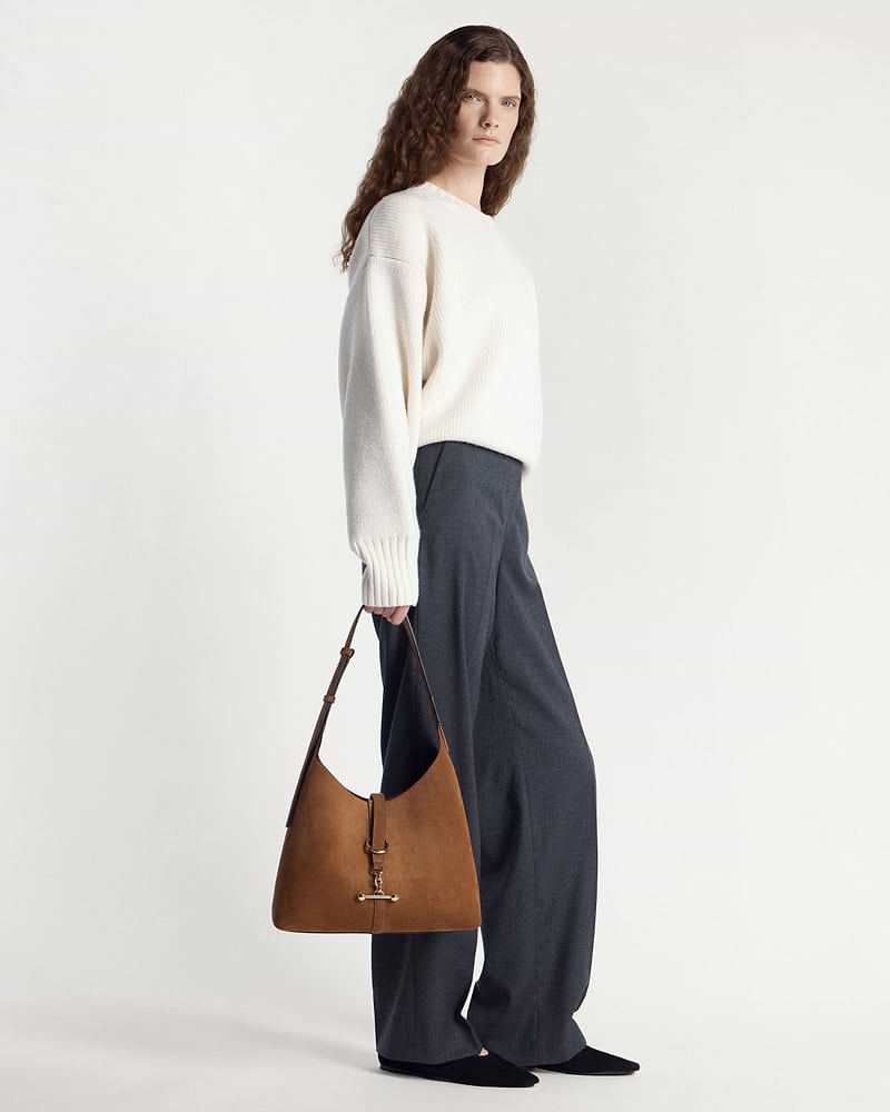 Model holding tan suede hobo bag with gold hardware at her side.
