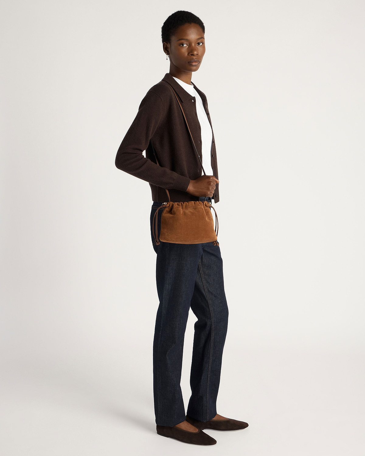 Model wearing tan suede drawstring bag across body with brown cardigan and dark jeans.