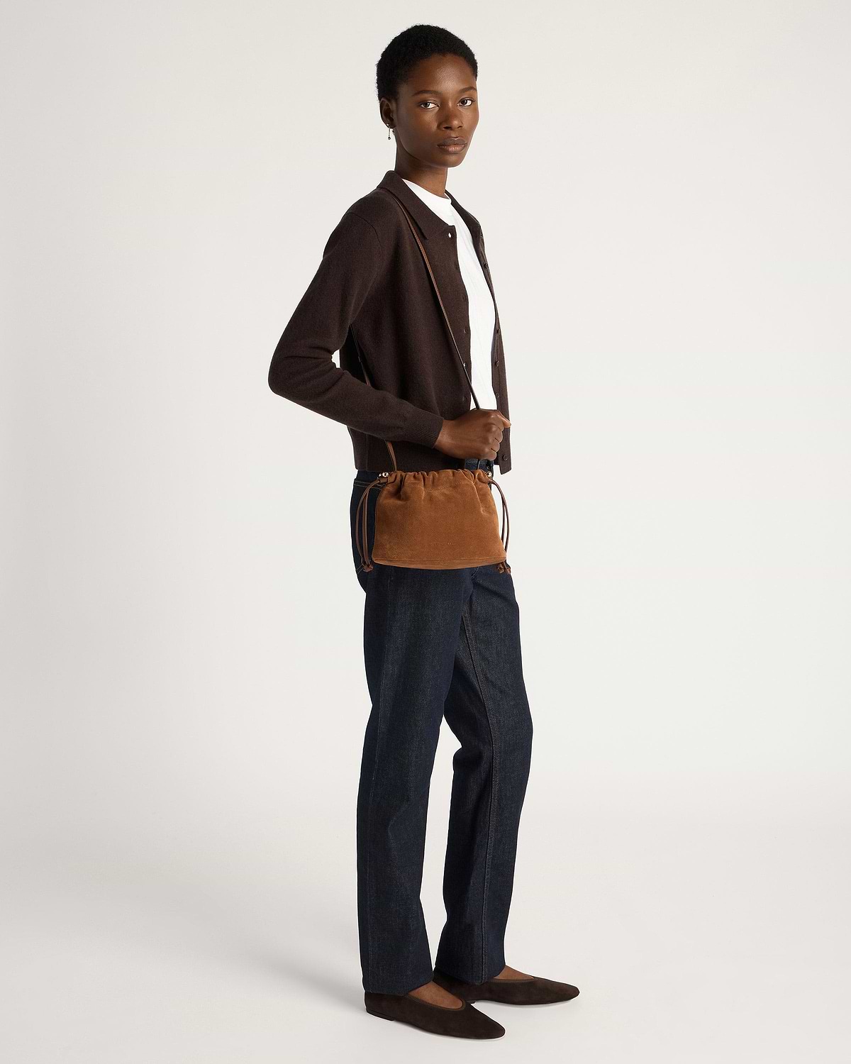 Model wearing tan suede drawstring bag across body with brown cardigan and dark jeans.