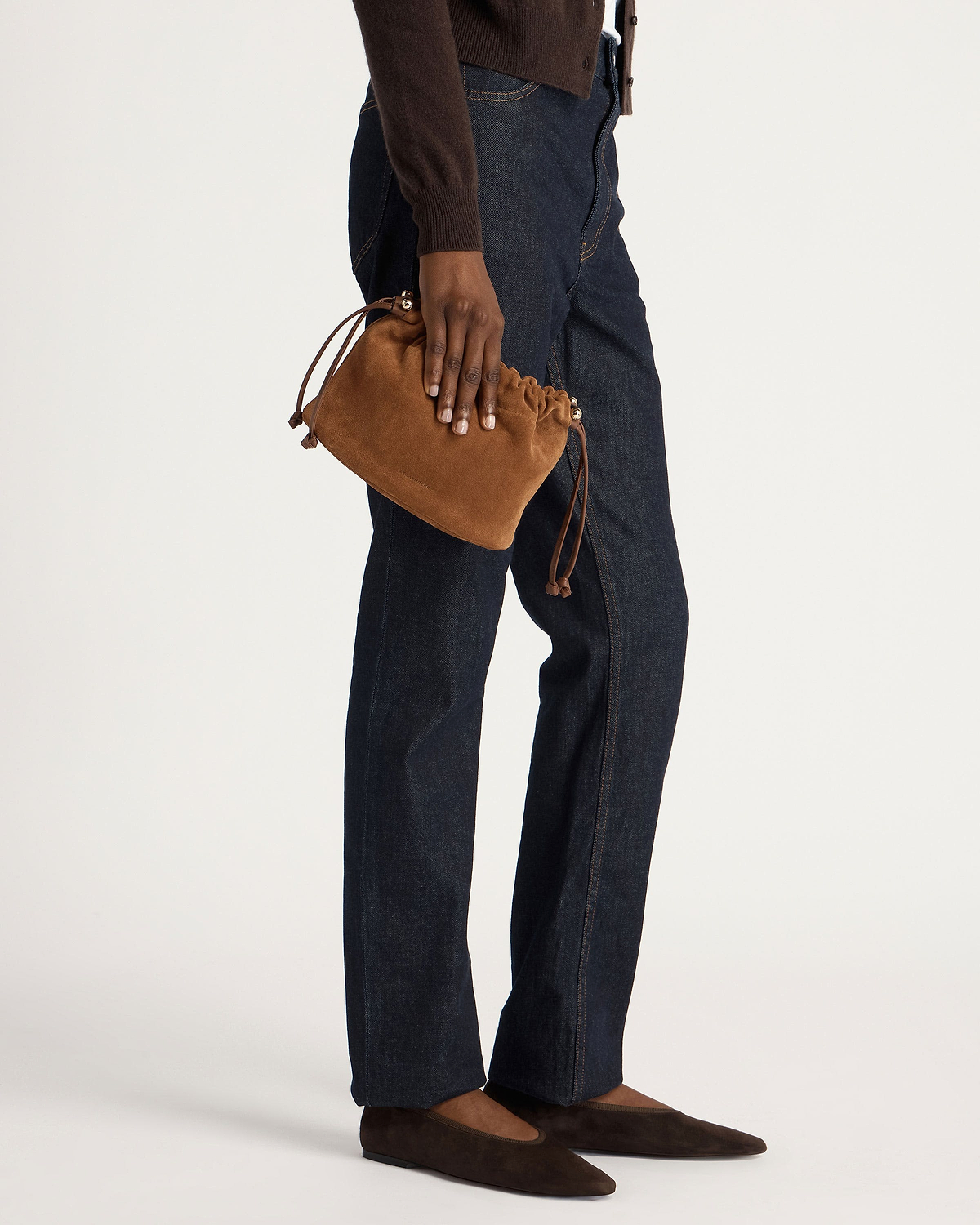 Tan suede drawstring bag held by model wearing dark jeans and brown flats.