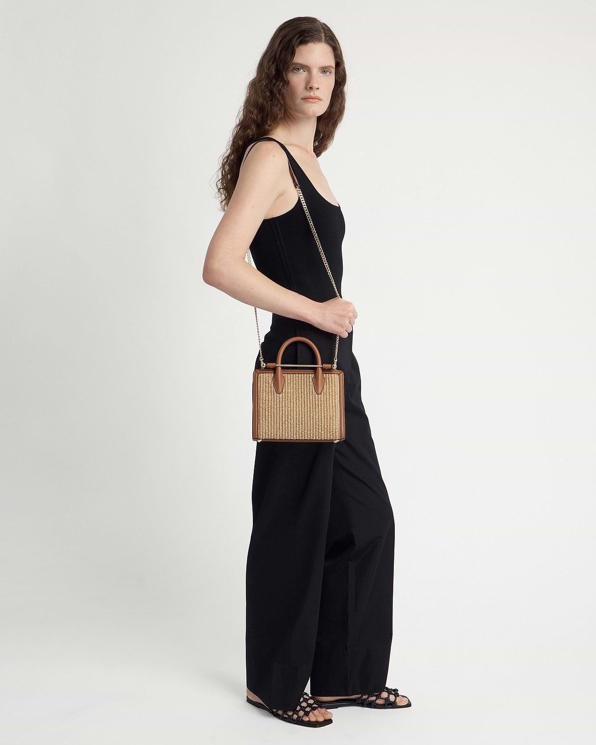 Woman in black outfit carrying tan raffia mini tote with leather trim and chain strap