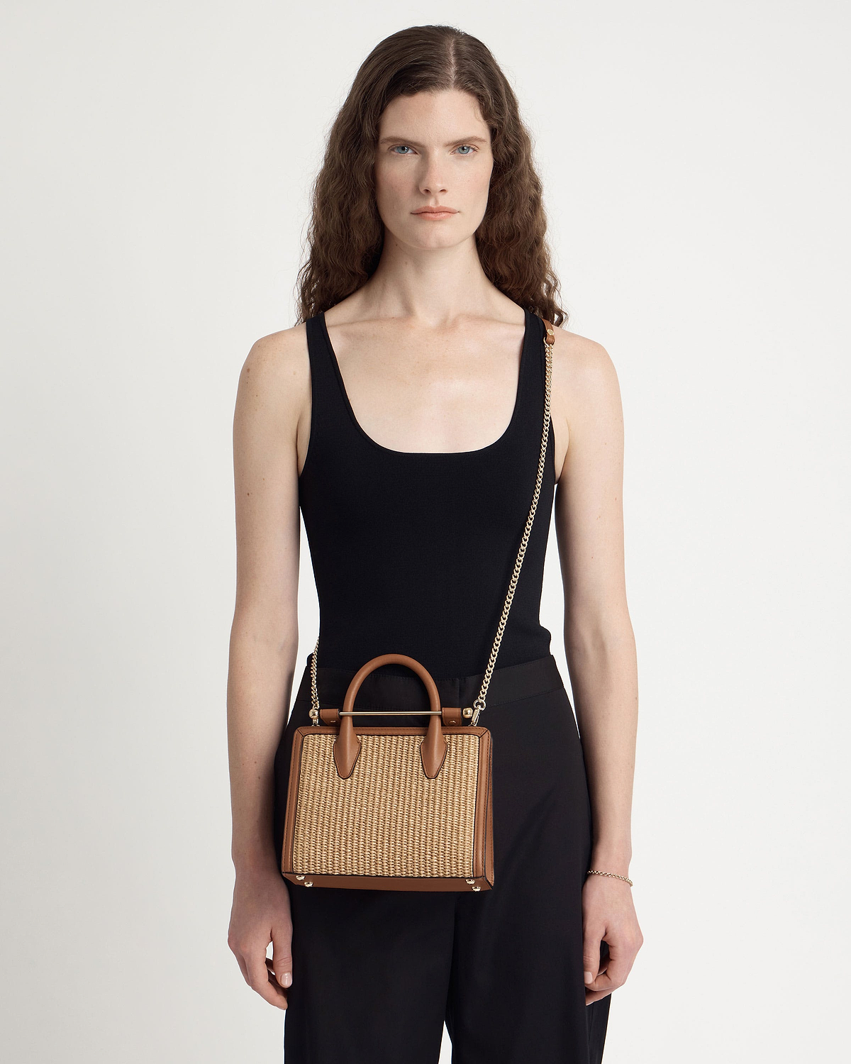 Model wearing black top holding tan and natural raffia mini tote with chain strap.