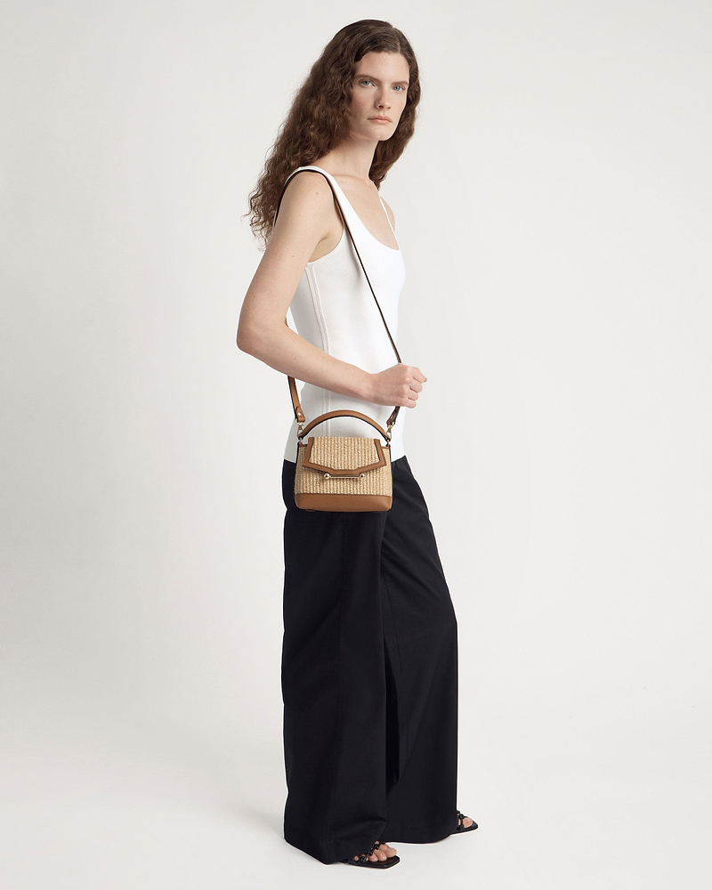 Model wearing white vest top and black wide trousers carrying tan raffia crossbody bag.