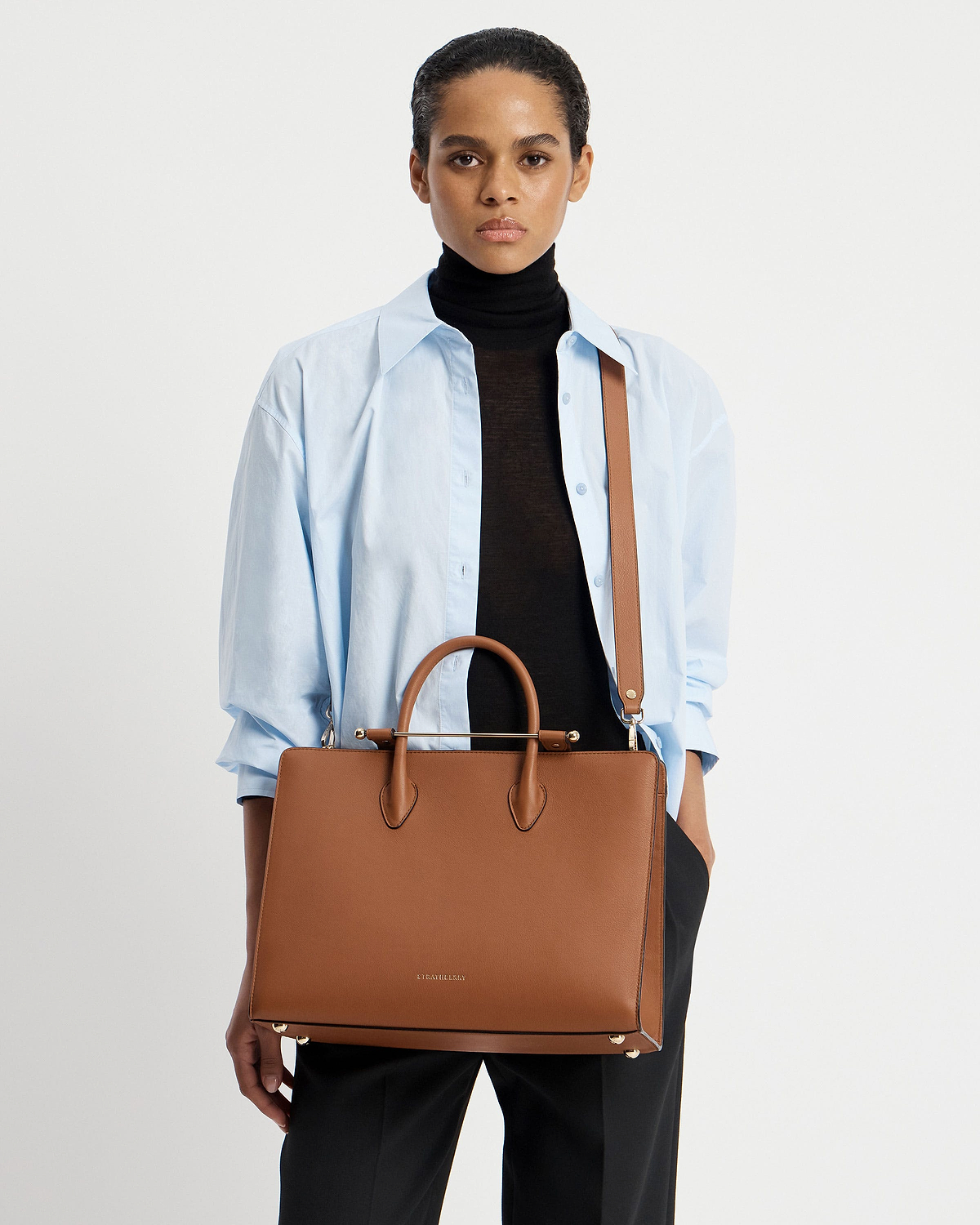 Model wearing light blue shirt and black turtleneck holding Strathberry chestnut tote.