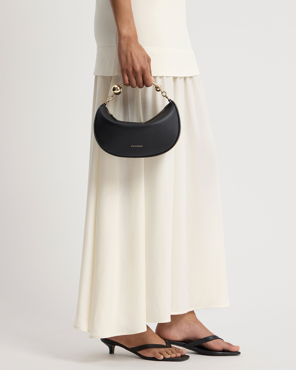 Handheld black Crescent Moon Mini handbag with gold ball handle, Strathberry logo.