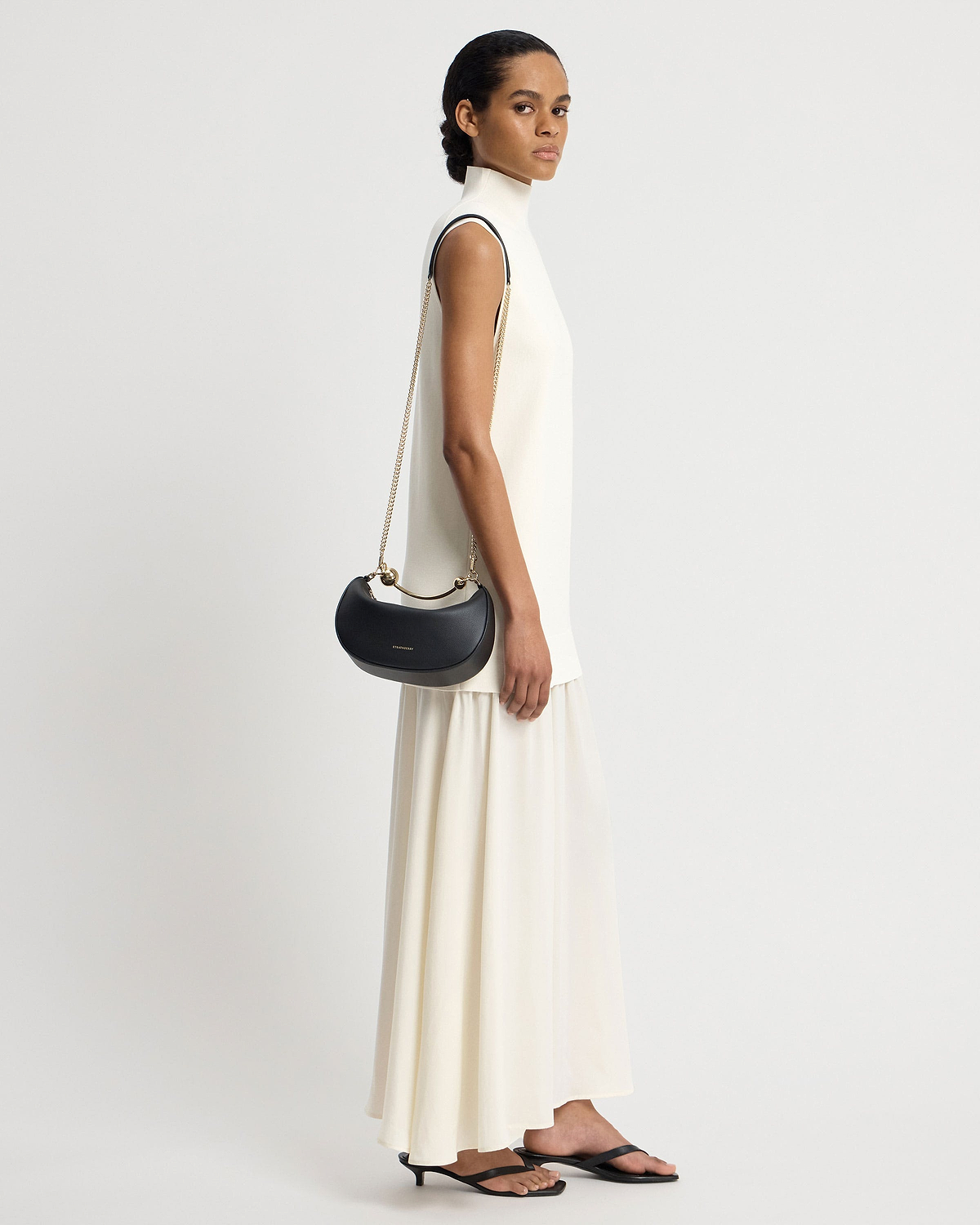 Model in cream dress carrying black crescent-shaped mini shoulder bag with gold chain.